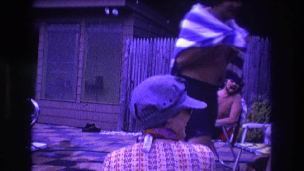 In 1975 Boston home-movie footage, a capped person in a patterned shirt faces away as a man dries off with a striped towel near a wooden fence and patio chairs.