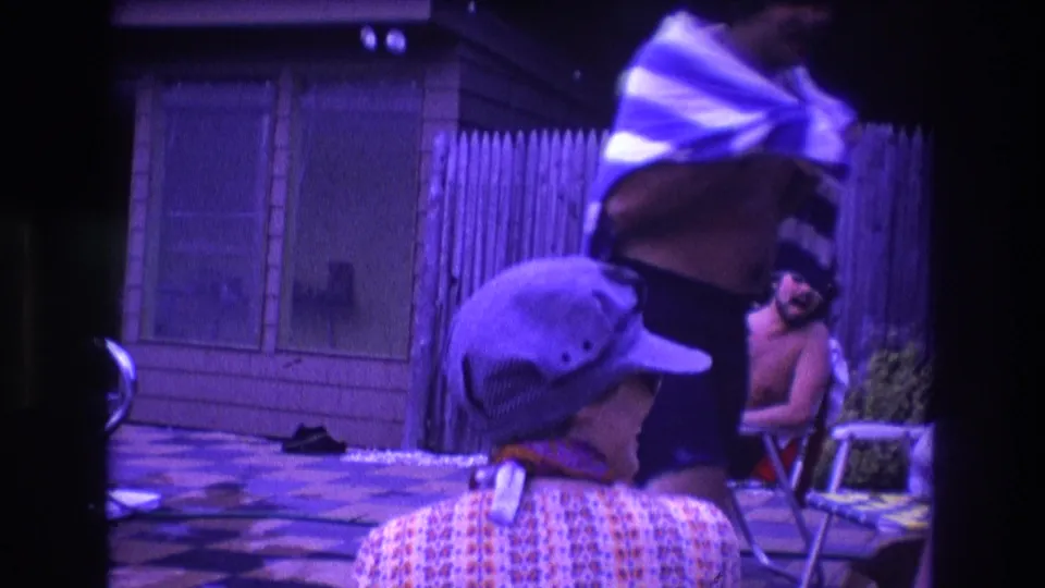 In 1975 Boston home-movie footage, a capped person in a patterned shirt faces away as a man dries off with a striped towel near a wooden fence and patio chairs.