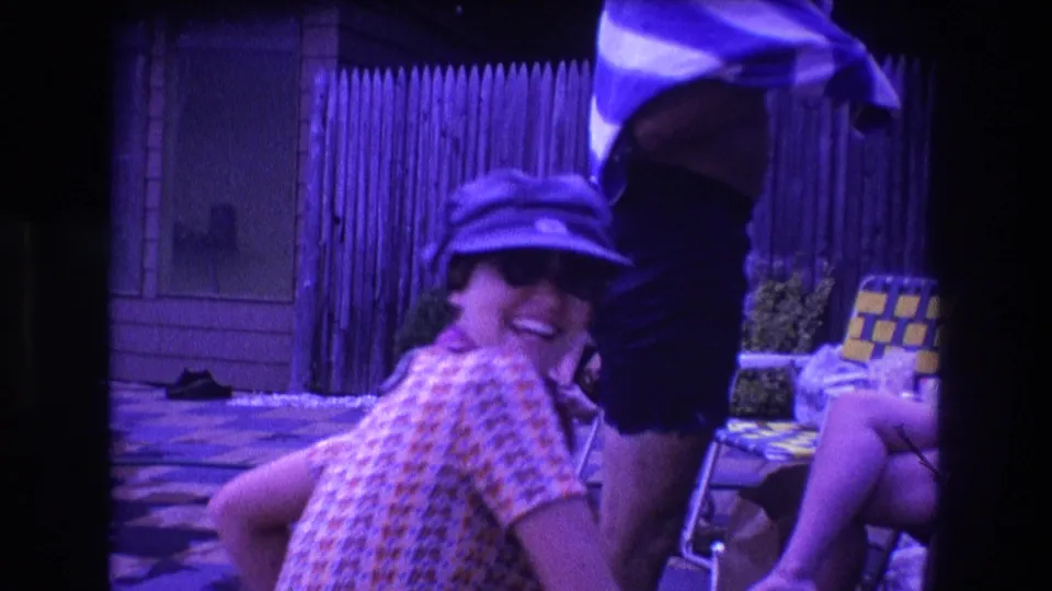 In the Boston 1975 home-movie frame, the capped person turns and smiles toward the camera beside a standing figure in swim trunks and a seated person on a folding chair.