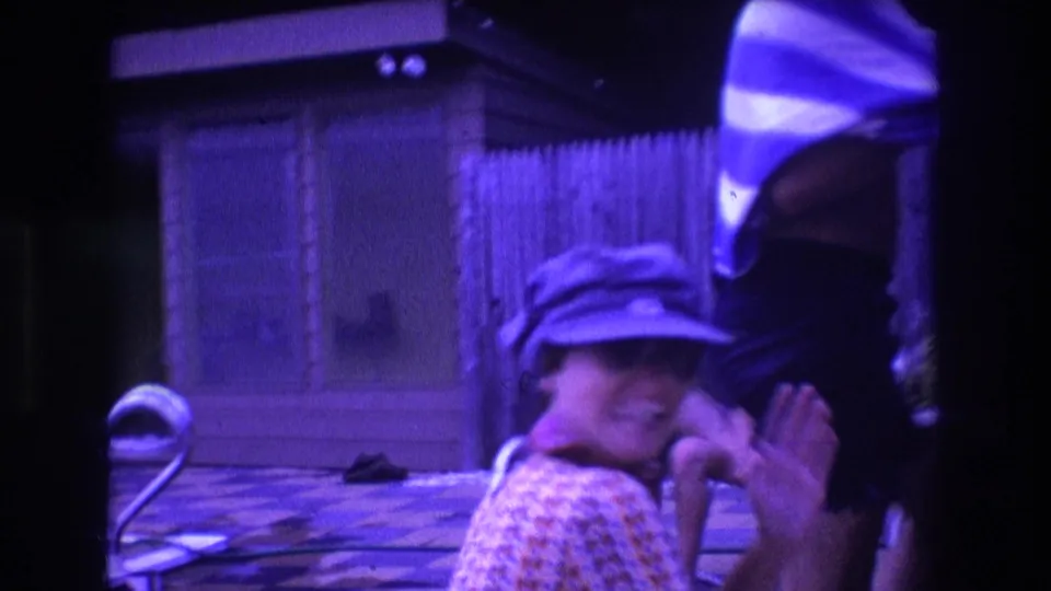 The final 1975 Boston frame catches the capped person laughing close to camera as a standing figure and backyard furniture remain in the purple-tinted 8mm scene.