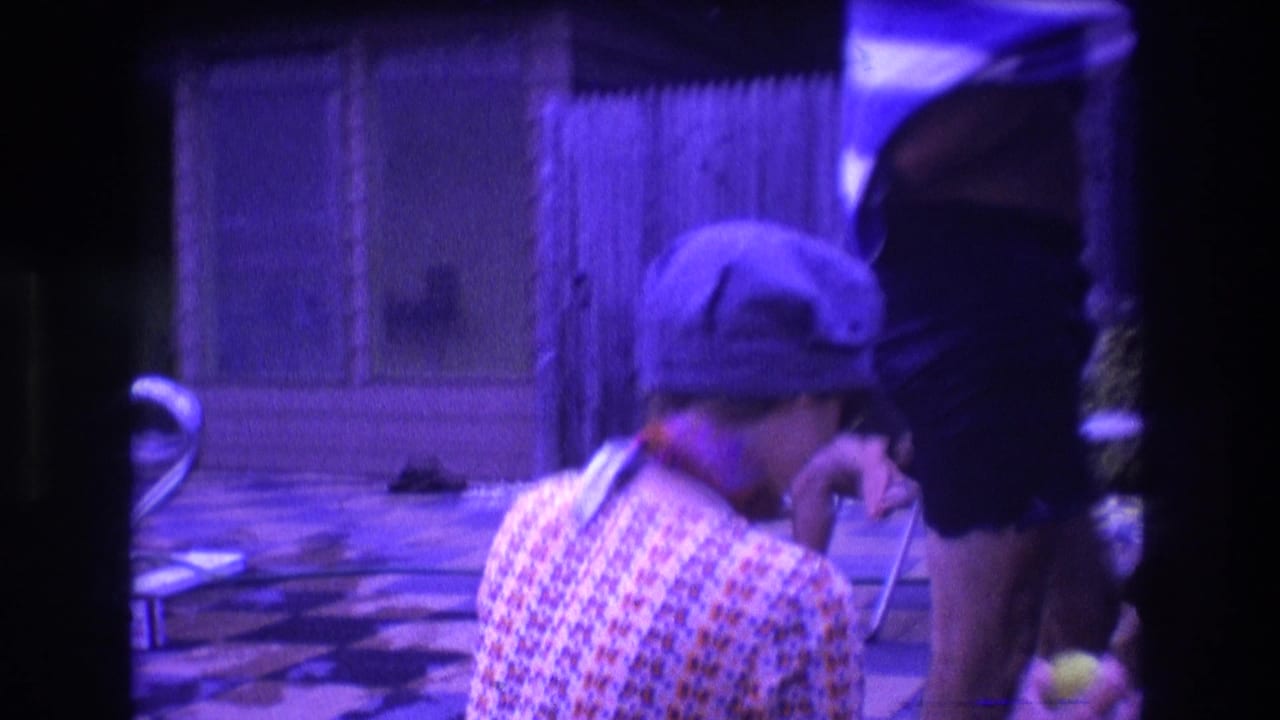 1975 Boston home-movie frame showing a woman in a blue cap and patterned shirt seated near a checker-pattern pool deck with adults and patio furniture nearby.