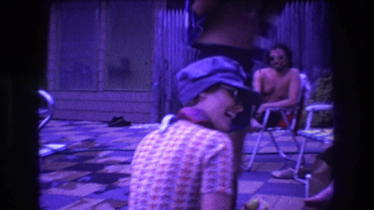 1975 Boston home-movie frame with a woman in a blue cap turning toward the camera on a backyard pool deck as adults sit in folding chairs behind her.