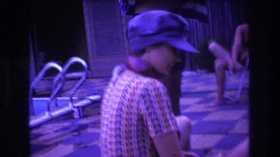 1975 Boston backyard pool frame showing a seated woman in a cap near a metal pool ladder, with adults and folding chairs in the softly focused background.