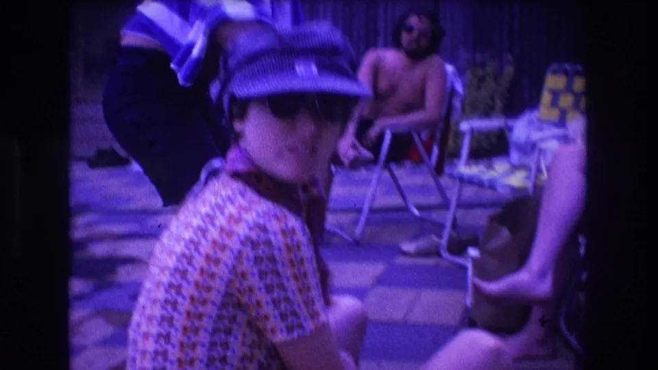 1975 Boston home-movie frame with a woman in a blue cap and sunglasses facing the camera amid seated and standing adults on a backyard pool deck.