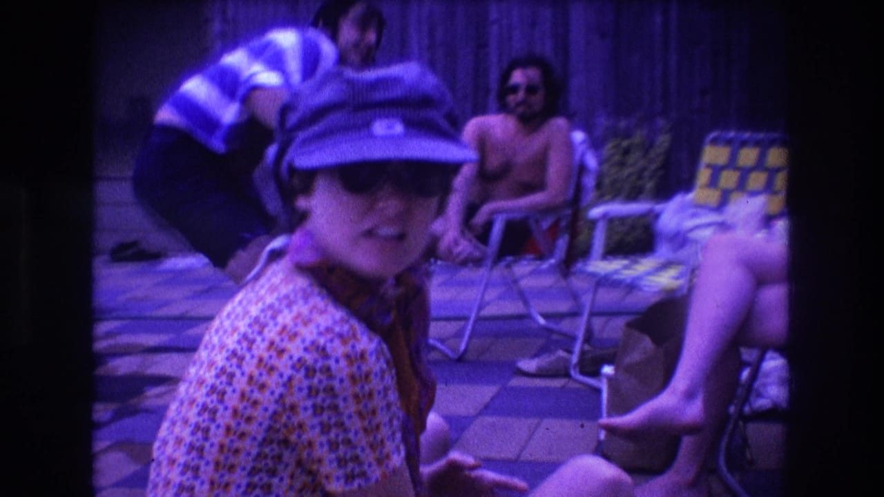 In a 1975 Boston 8mm home-movie frame, a woman in a patterned dress, blue cap, sunglasses, and neck scarf faces the camera near a backyard pool while adults lounge behind her.