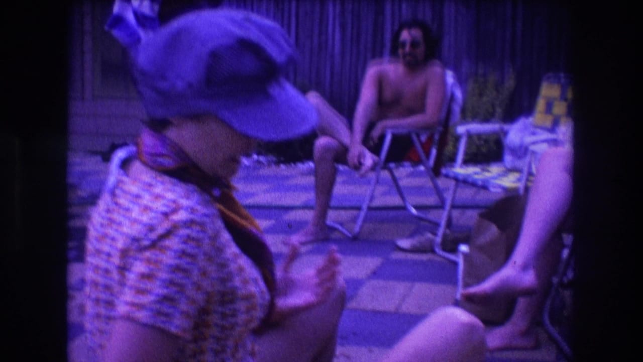 The woman in the patterned dress and blue cap turns in profile near patio chairs, with a shirtless man and another seated adult visible in the purple-tinted 8mm frame.