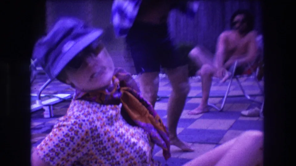 A woman in sunglasses, cap, patterned dress, and scarf leans back close to the camera while adults pass or sit around the poolside patio in an informal home-movie moment.