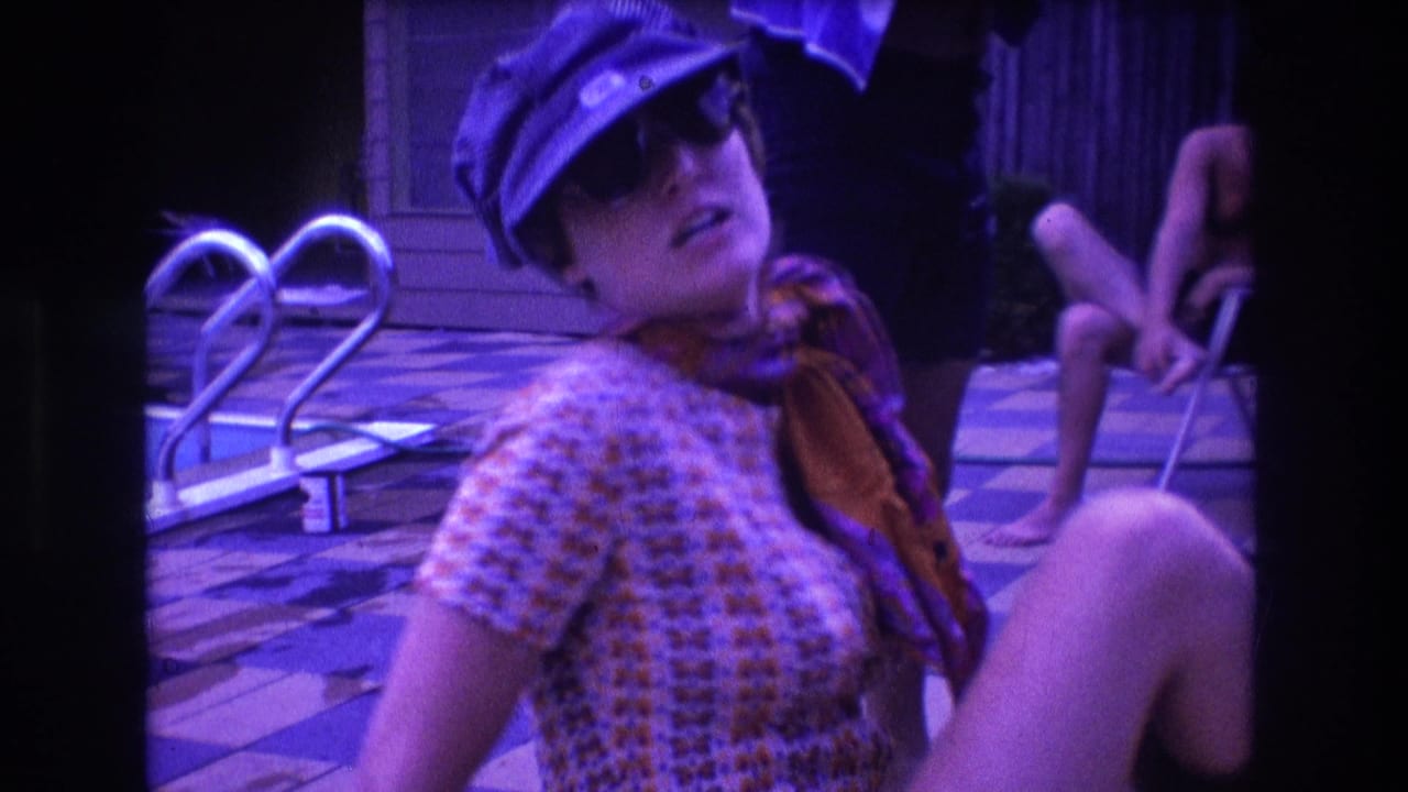 In 1975 Boston home-movie footage, a woman wearing sunglasses, a blue cap, patterned dress, and colorful scarf sits near a pool while a shirtless man is seated in the background.