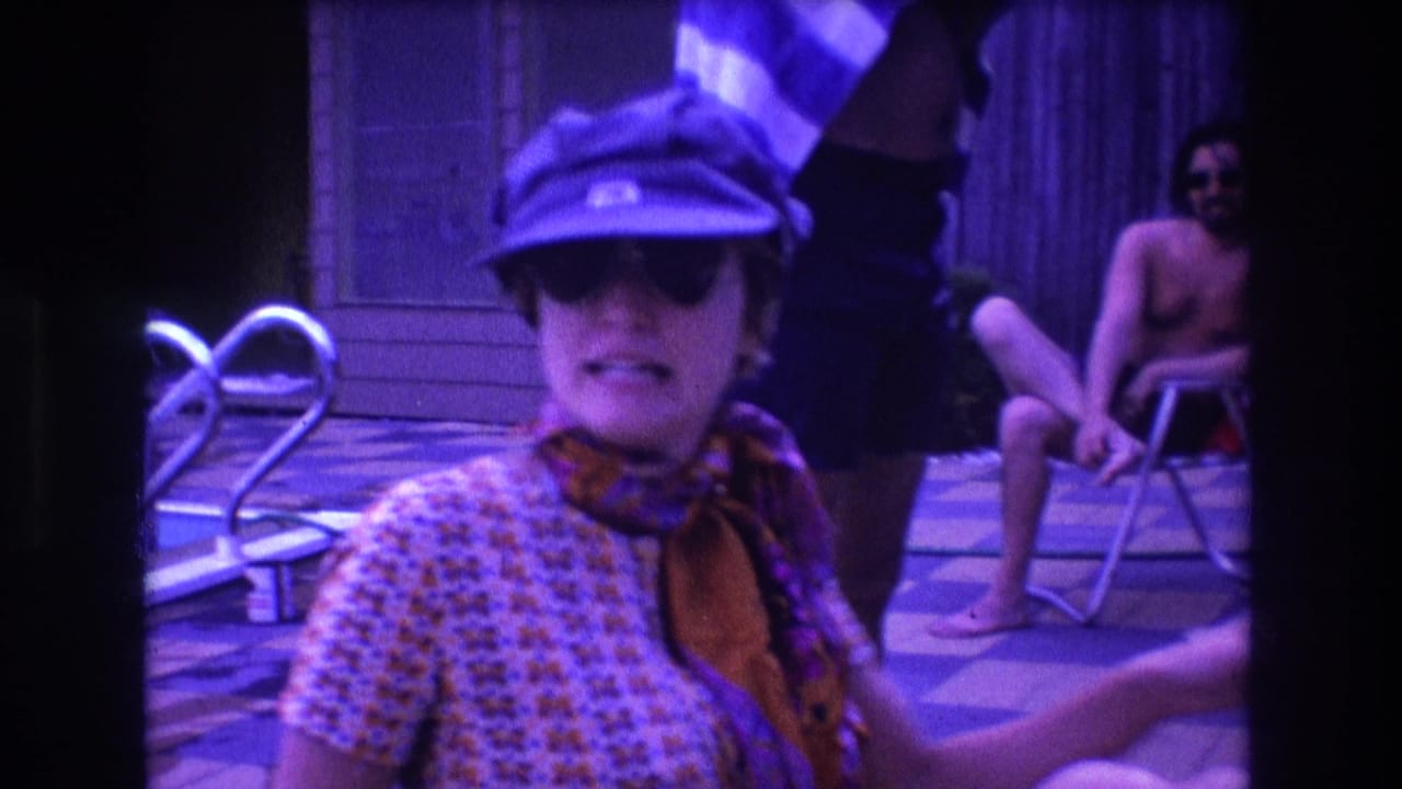 The woman faces the camera in a purple-tinted 1975 Boston 8mm poolside scene, with a seated shirtless man and a blue-and-white striped towel visible behind her.