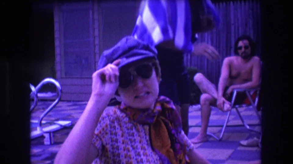 The woman lifts a hand to her sunglasses or cap in a candid 1975 Boston poolside home-movie frame, while a shirtless man sits in a folding chair behind her.