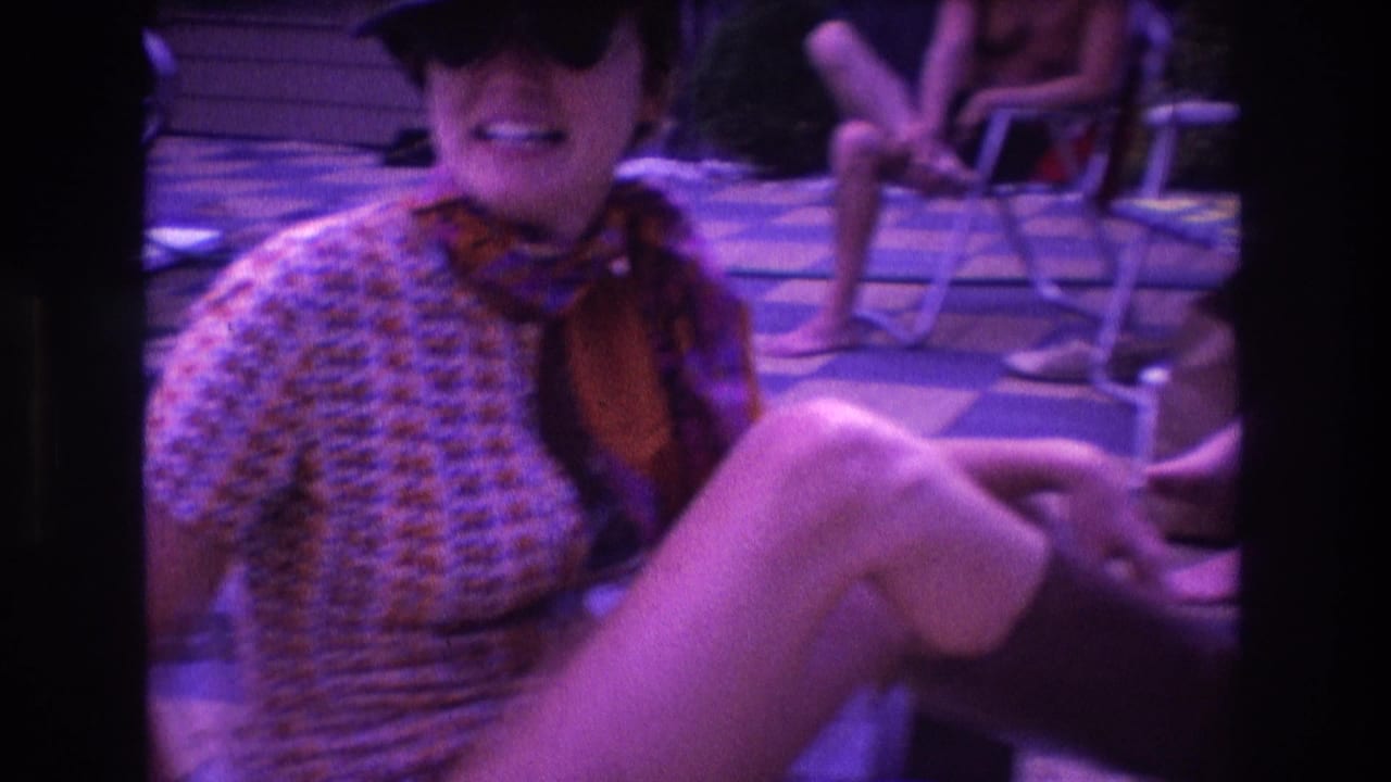 In 1975 Boston home-movie footage, a woman in a hat and patterned shirt sits on a folding patio chair and raises a tall brown boot while other people lounge behind her.