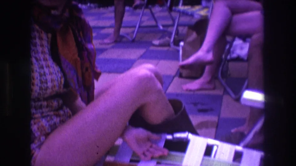 1975 Boston home-movie frame showing a seated woman extending her leg beside a folding lawn chair, with friends' legs and bare feet visible in the surrounding patio scene.