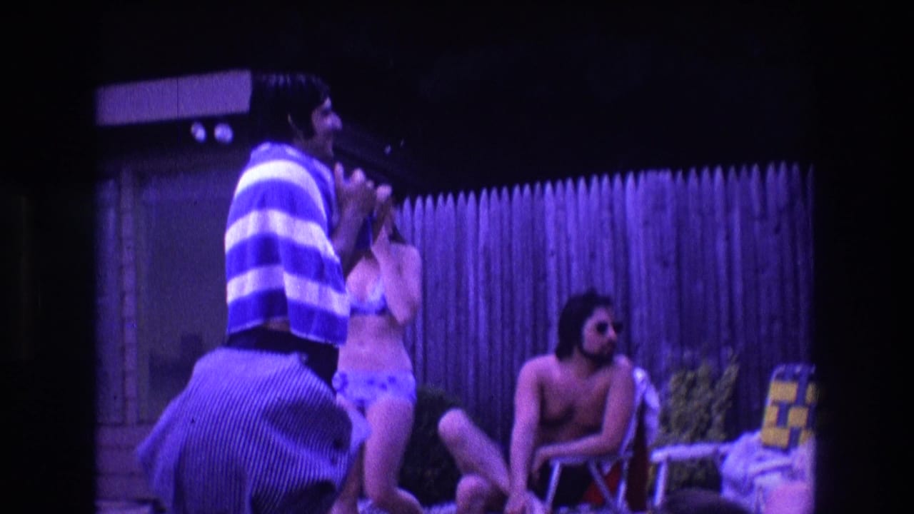 A man wrapped in a striped towel gestures while adults in swimwear sit and stand near a fence.