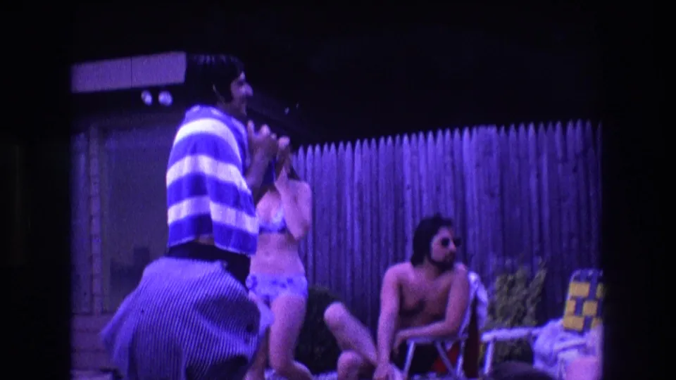 1975 Boston home-movie frame with a man in a blue-and-white striped towel gesturing near adults in swimwear at a backyard pool gathering.