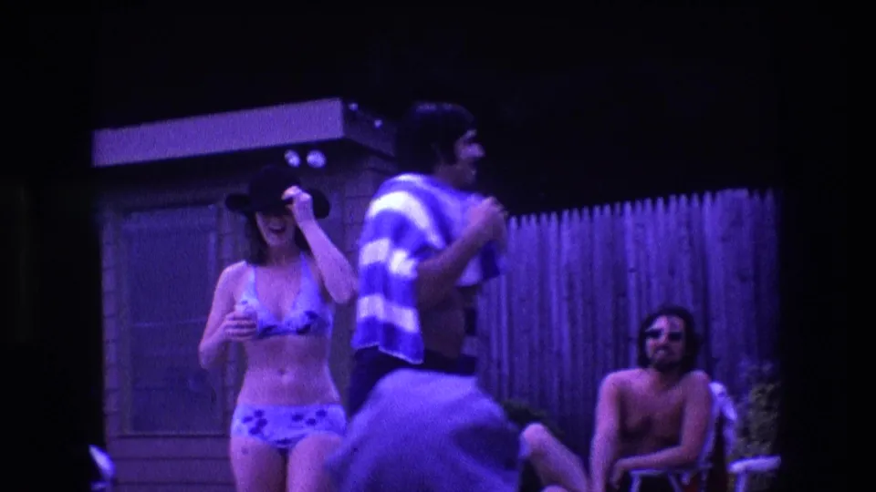 1975 Boston 8mm frame showing a woman in a bikini and cowboy hat holding a drink while a man in a striped towel stands near the wooden fence.