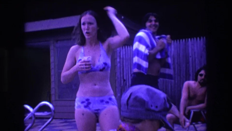 1975 Boston backyard pool party frame with a woman in a bikini holding a drink in the foreground, pool railings, a striped towel, and seated adults behind her.