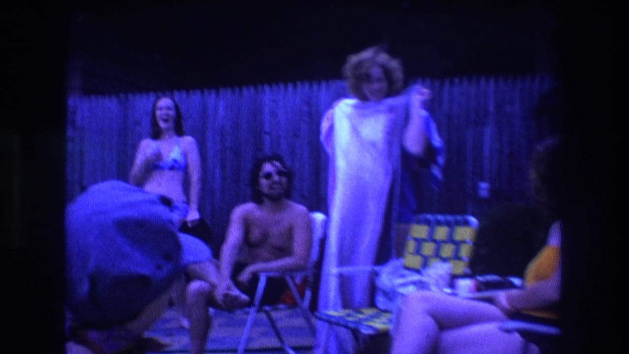 1975 Boston 8mm home-movie frame showing young adults in a backyard, including a seated shirtless man, a woman in swimwear, and another woman with a towel near folding chairs.