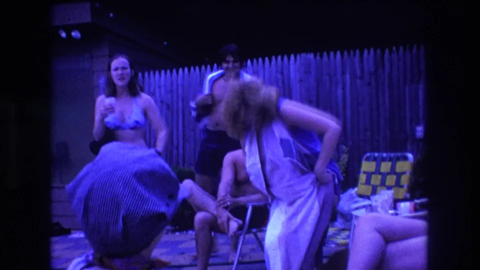 1975 Boston home-movie frame showing young adults in a backyard, with a woman bending while wrapped in a towel, a woman in swimwear, a standing man, and a seated man near folding chairs.