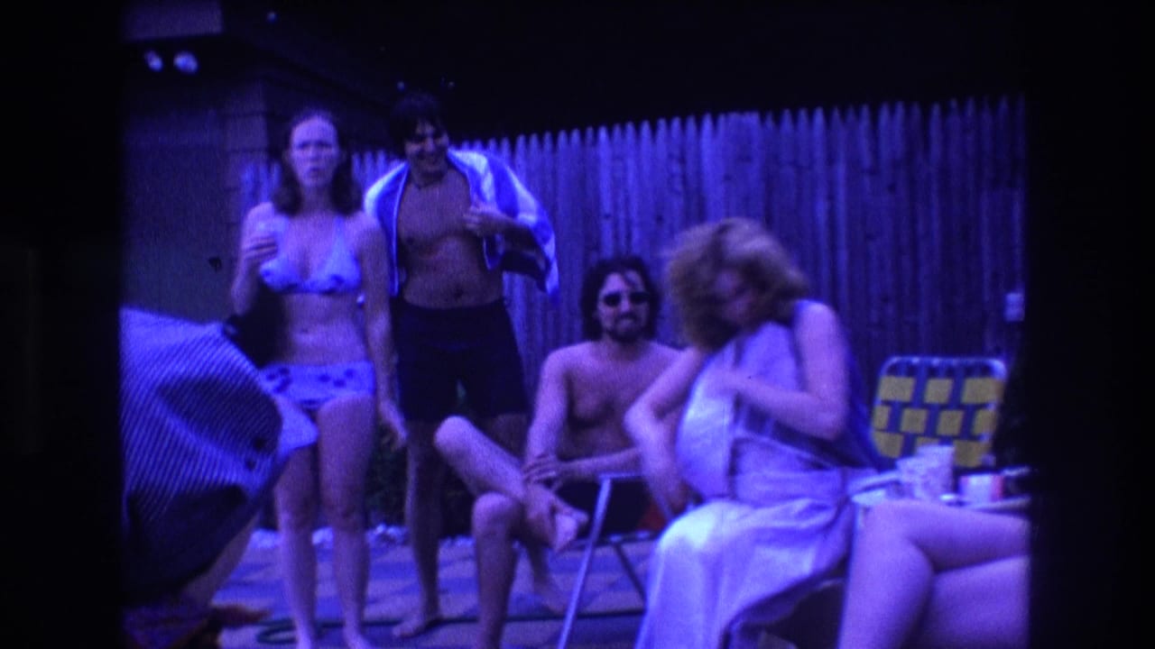 1975 Boston home-movie frame showing adults at a backyard pool party, with one person holding a drink, another wrapped in a towel, and others seated near folding lawn chairs.