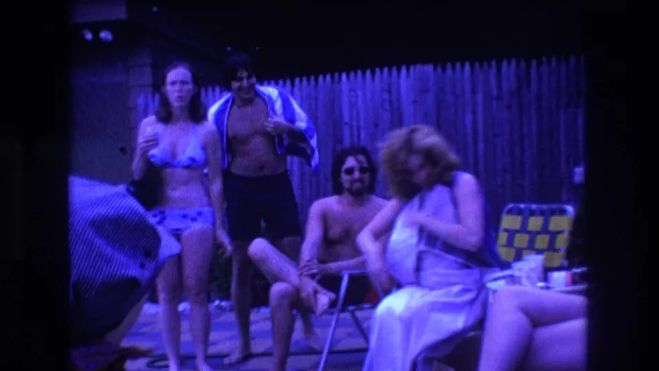 1975 Boston home-movie frame showing adults at a backyard pool party, with one person holding a drink, another wrapped in a towel, and others seated near folding lawn chairs.