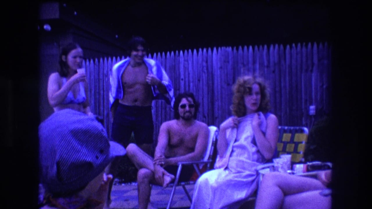 1975 Boston home-movie frame of adults relaxing near a wooden backyard fence, including a seated shirtless man in sunglasses and a woman in a light swimsuit holding a drink.