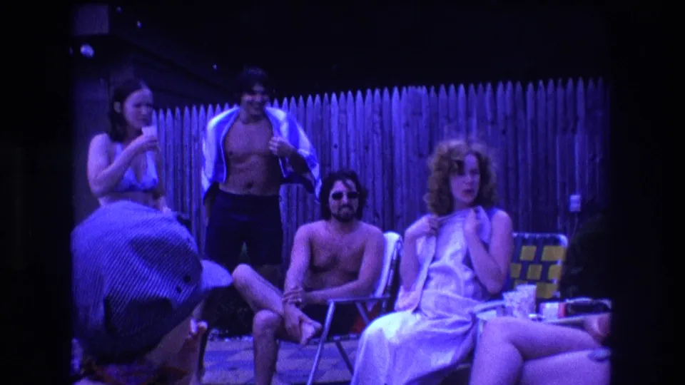 1975 Boston home-movie frame of adults relaxing near a wooden backyard fence, including a seated shirtless man in sunglasses and a woman in a light swimsuit holding a drink.