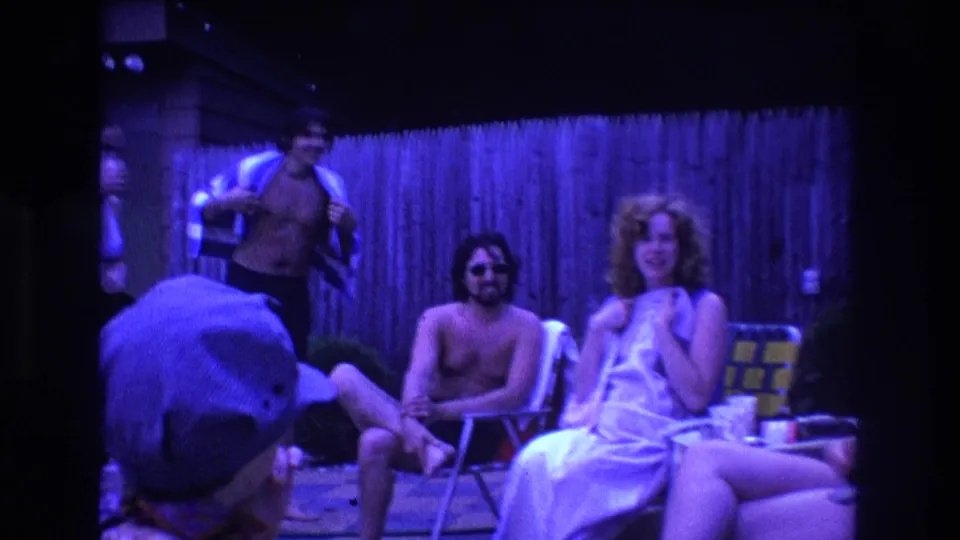 1975 Boston home-movie frame showing a candid backyard party moment, with adults in swimwear near folding chairs and a foreground figure partially blocking the view.