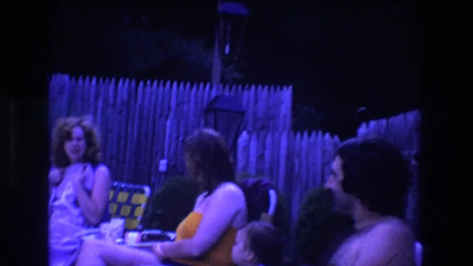 1975 Boston home-movie frame showing adults seated together at a backyard pool party, with a wooden fence behind them and dim blue-purple night lighting across the scene.