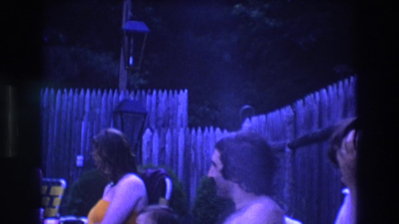 In a 1975 Boston 8mm home-movie frame, several adults sit close together in a dim backyard with a wooden picket fence and strong blue-purple color cast.