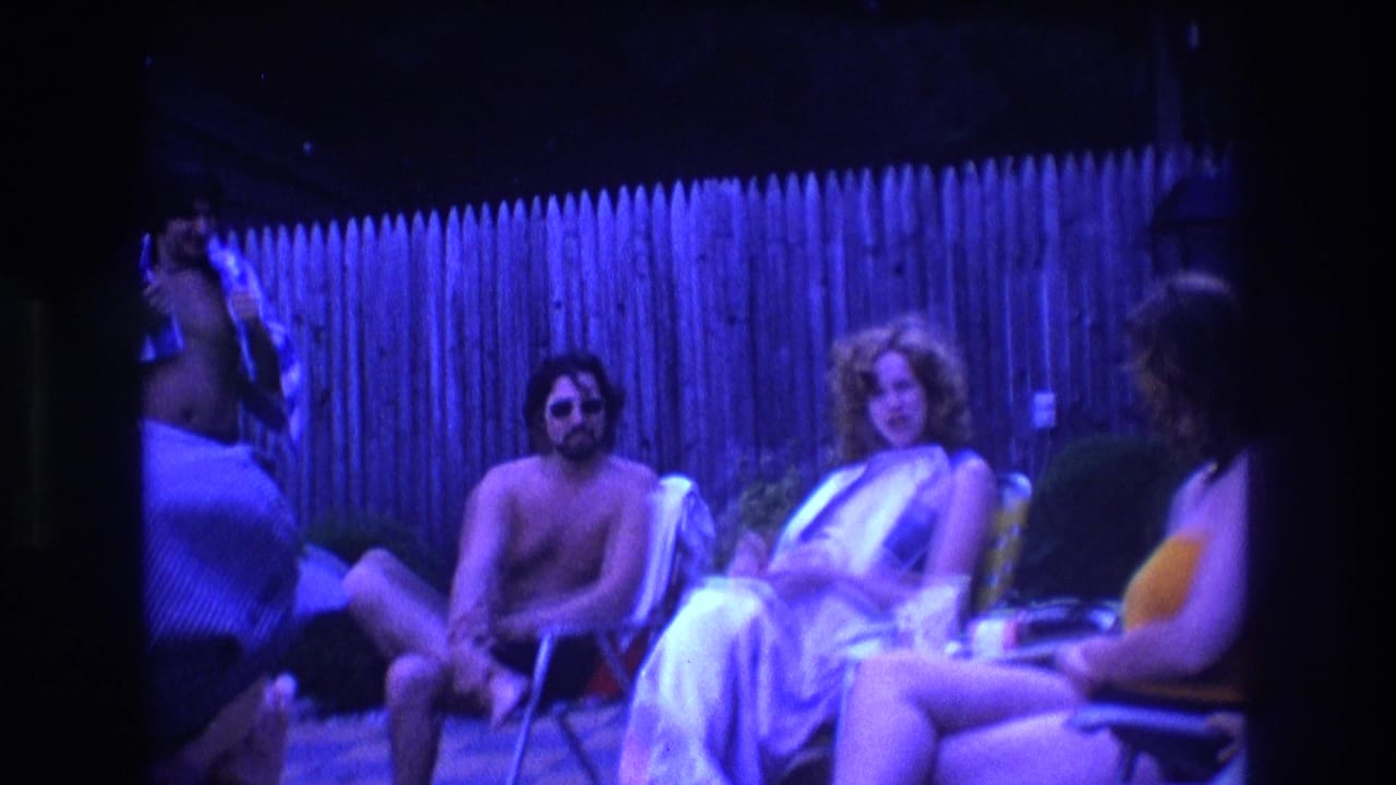 Adults sit on folding chairs near a picket fence, including a shirtless man wearing sunglasses.