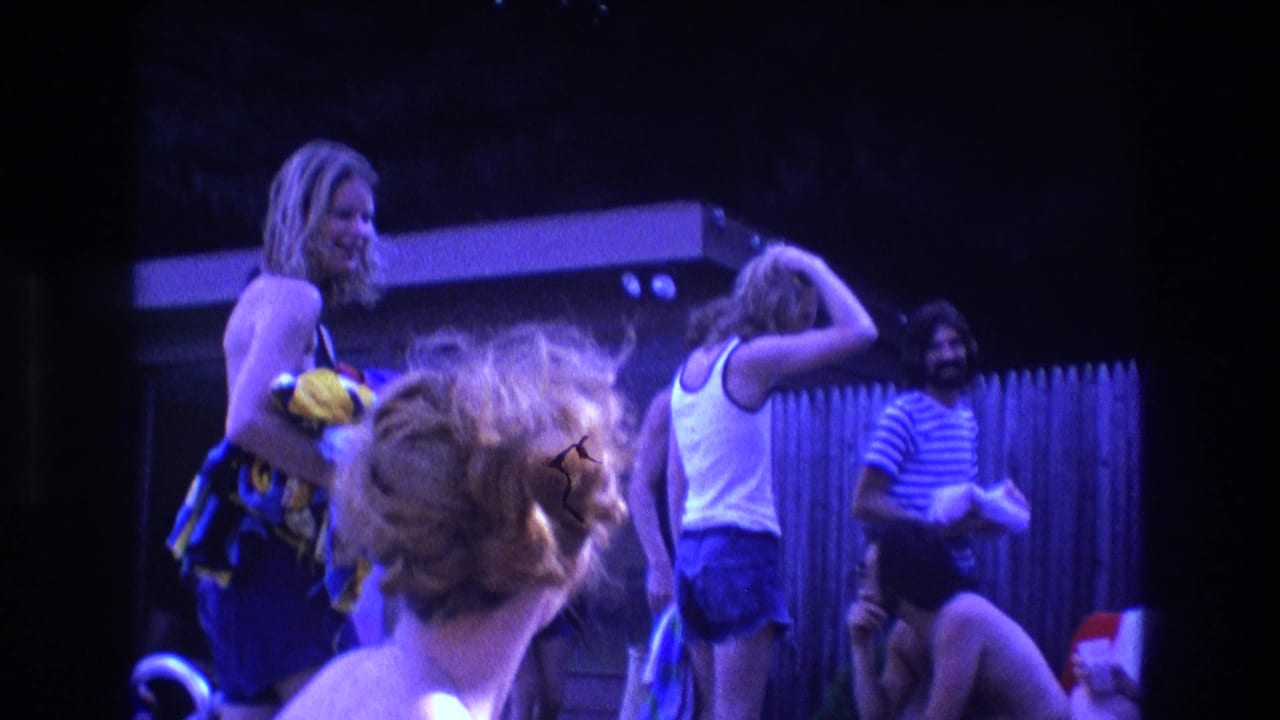 1975 Boston 8mm frame showing young adults at a backyard pool gathering, with a woman holding a yellow bag and others standing or seated near a wooden fence.
