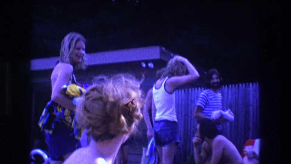 1975 Boston 8mm frame showing young adults at a backyard pool gathering, with a woman holding a yellow bag and others standing or seated near a wooden fence.
