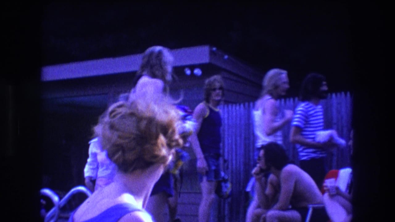 1975 Boston home-movie frame of young adults in tank tops and swimsuits chatting by a backyard fence as the gathering begins to break up.