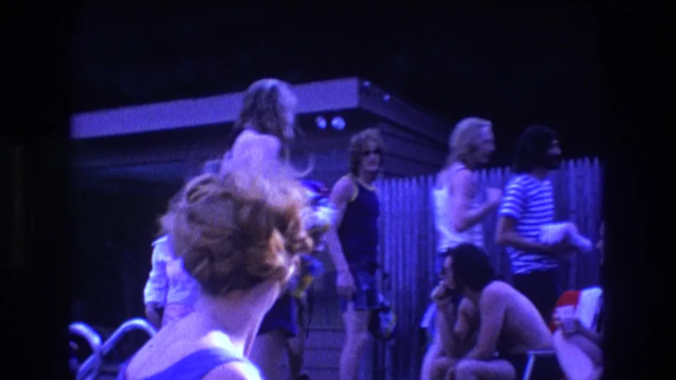 1975 Boston home-movie frame of young adults in tank tops and swimsuits chatting by a backyard fence as the gathering begins to break up.