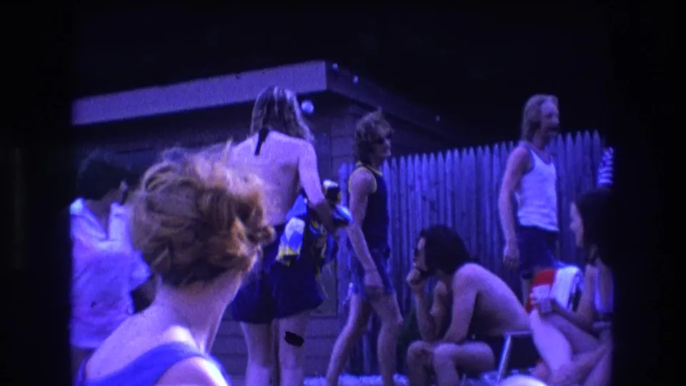 1975 Boston 8mm frame showing a candid poolside goodbye moment, with people in summer clothing gathered near a fence, shed, and lounge chairs.