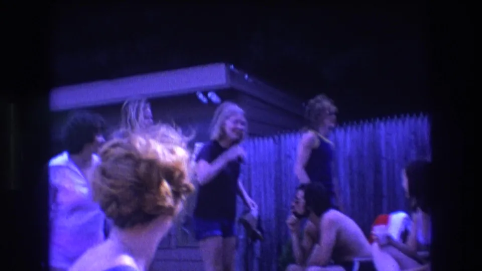 1975 Boston 8mm frame of pool party guests in motion near a backyard fence, with soft focus and blue-purple evening color from the film scan.