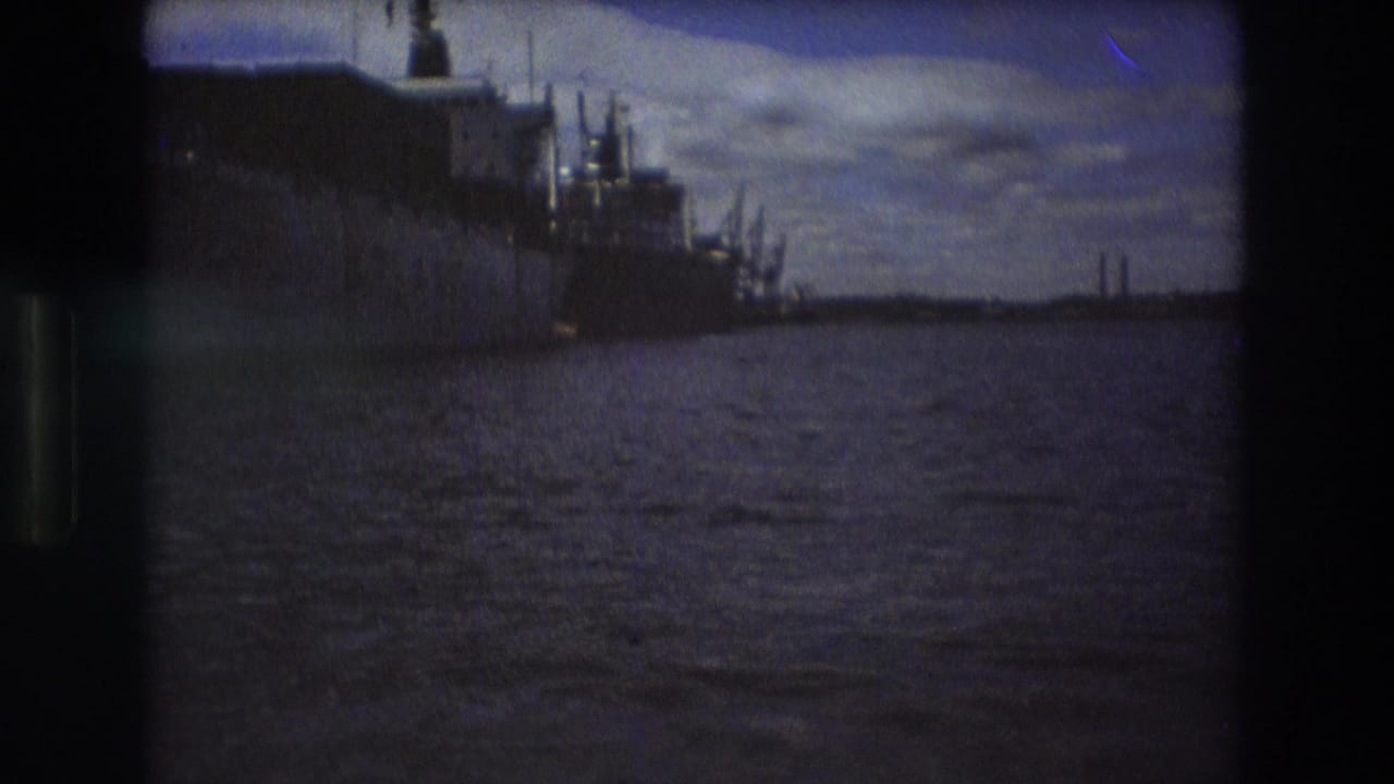 Boston harbor in the supplied 1975 metadata, with distant docked ships, rippled water, cloudy sky, and dark film-frame borders from the 8mm scan.
