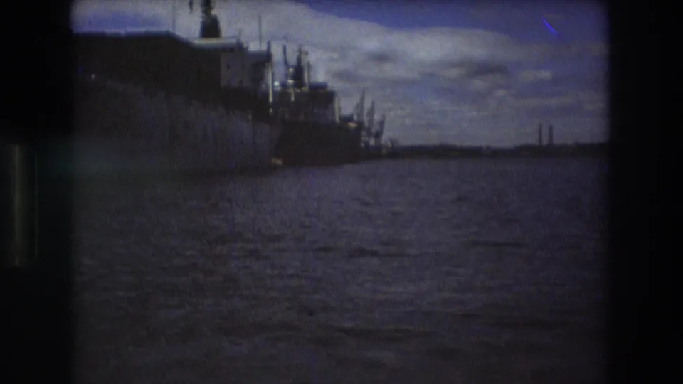 Boston harbor in the supplied 1975 metadata, with distant docked ships, rippled water, cloudy sky, and dark film-frame borders from the 8mm scan.
