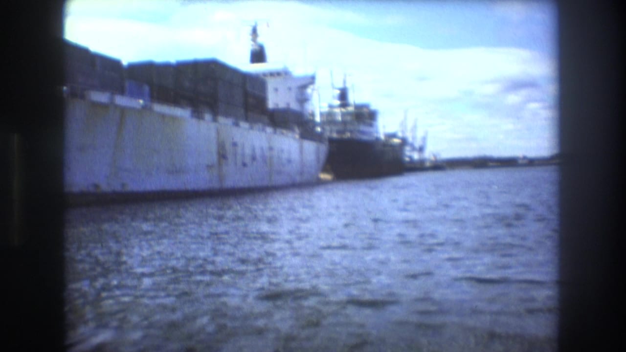 Large docked cargo ship beside harbor water with another vessel behind it.