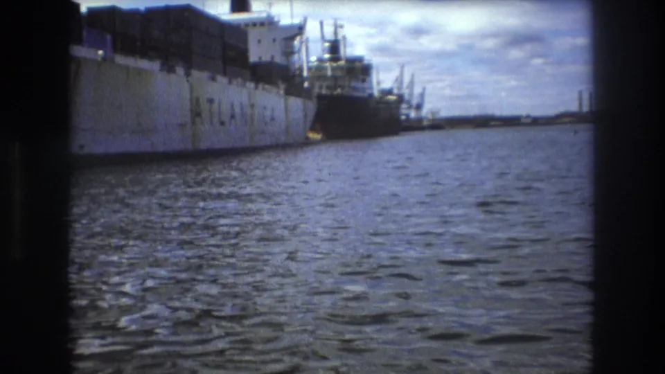 Side view of the docked cargo ship in the supplied 1975 Boston harbor clip, with rippled water, a dark vessel behind, and cranes or masts near the dock.