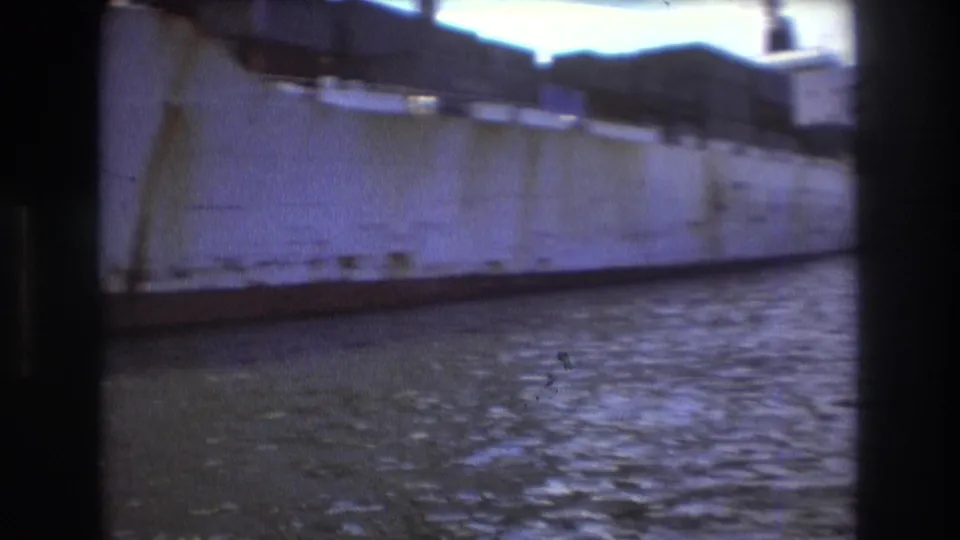 Close moving view along the pale hull of the cargo ship in the Boston harbor footage, with water ripples below and soft 8mm blur at the frame edges.