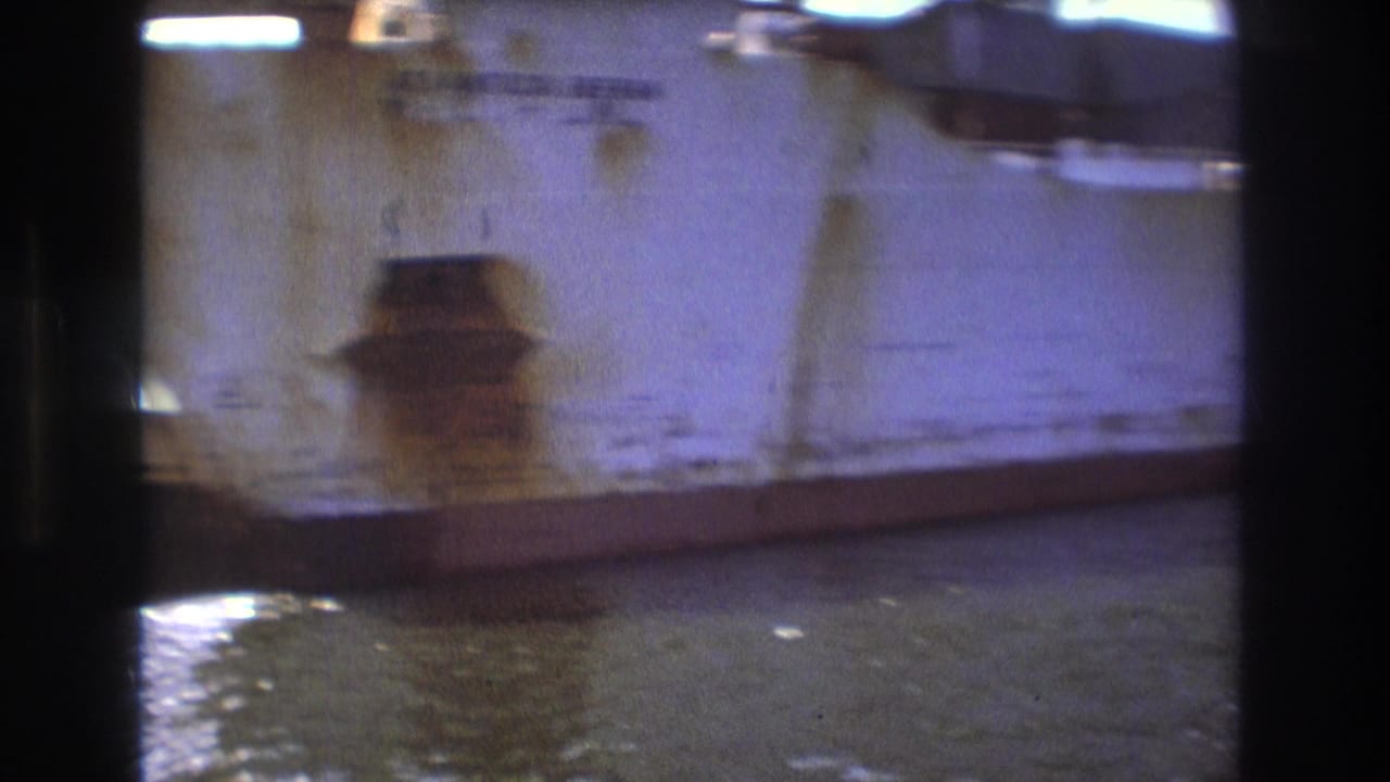 A weathered cargo ship hull appears close to the camera in Boston harbor, with rust marks, dark film-frame borders, and water below.