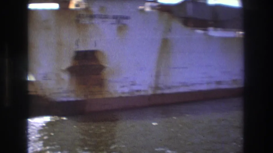 A weathered cargo ship hull appears close to the camera in Boston harbor, with rust marks, dark film-frame borders, and water below.