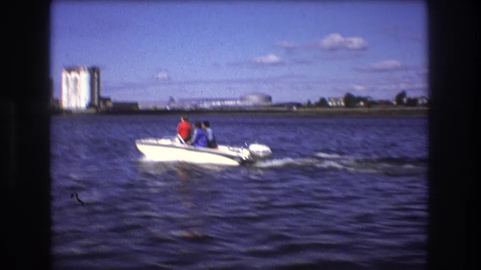 Two people in a small white speedboat pass across Boston harbor, with choppy water, industrial buildings, storage tanks, and a low shoreline in the background.