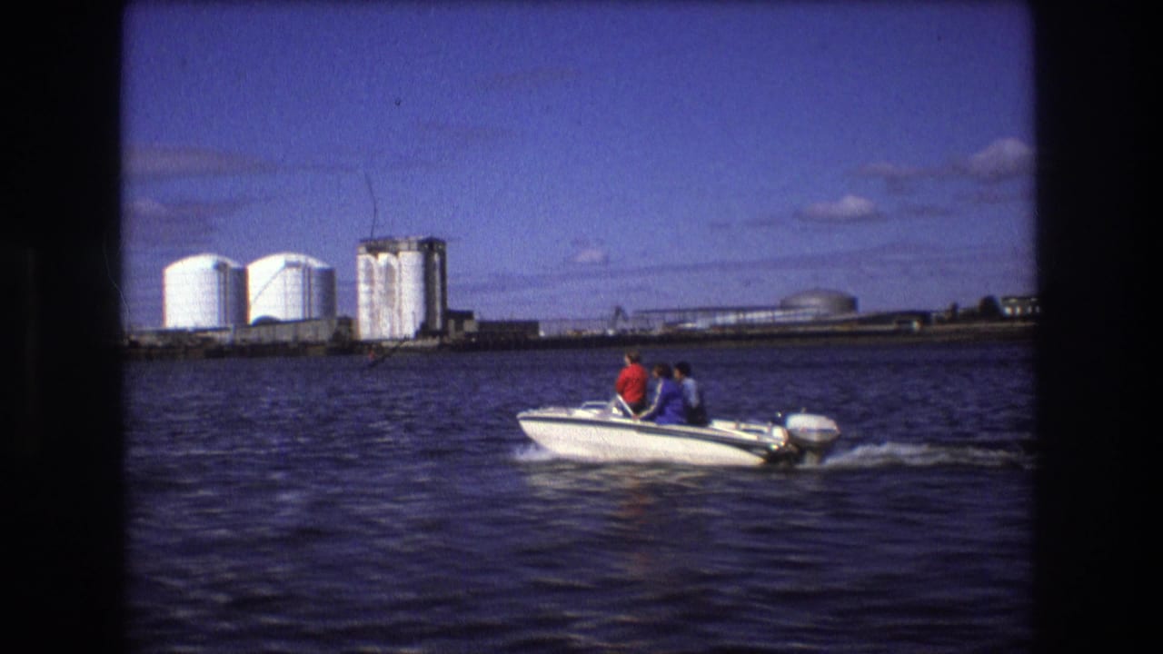 In supplied 1975 Boston home-movie footage, two seated people ride a small white outboard motorboat across dark river water with white storage tanks and industrial buildings in the background.