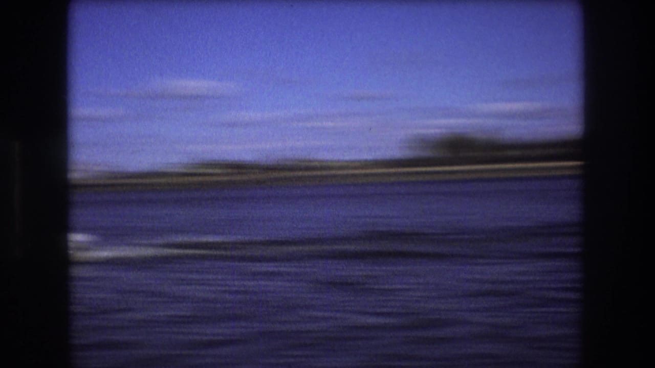 A motion-blurred river view from the moving boat shows dark water, a low distant shoreline, and open blue sky in the supplied Boston 1975 footage.