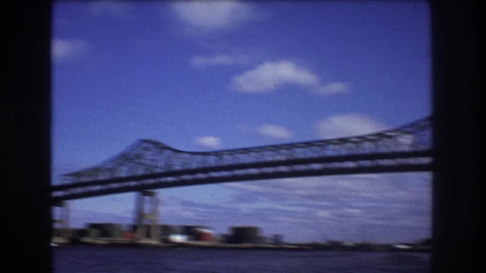 The moving 8mm view passes alongside a large steel truss bridge over the river, with low waterfront structures and blue sky visible below and behind it.