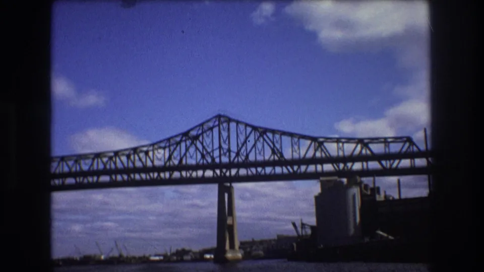 In the supplied Boston 1975 clip, a large steel truss bridge crosses the river under a blue sky, with industrial tanks and waterfront structures visible near the right side of the frame.
