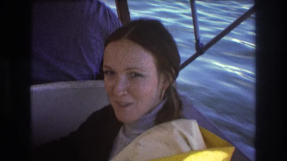 A young brunette woman in a turtleneck and life vest looks toward the camera from a small motorboat, with moving blue water behind her.