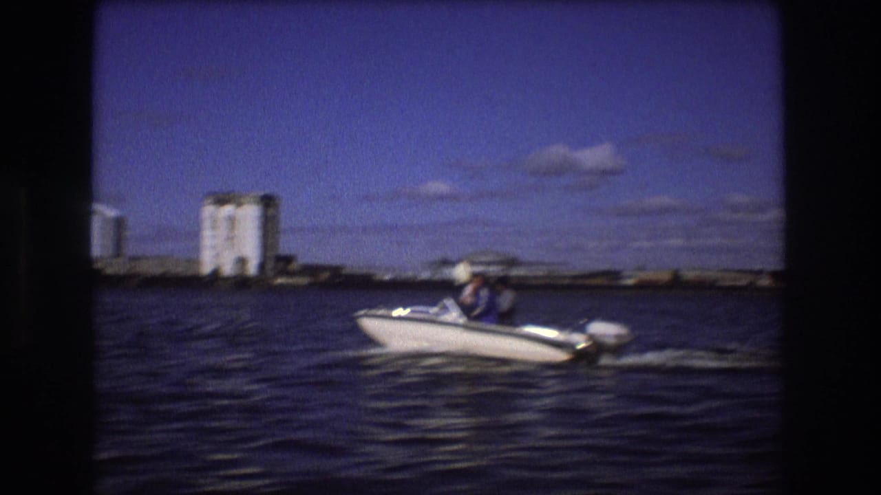 1975 Boston home-movie frame showing a white outboard motorboat with visible passengers moving across harbor water, with industrial shoreline structures in the distance.